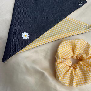 Könnte beinhalten: Ein dreieckiges Halstuch mit einer Seite aus dunkelblauem Denim und einer Seite mit gelb-weißem Vichy-Karo-Muster, verziert mit einer weißen Gänseblümchen-Stickerei und Metalldruckknöpfen, dazu ein passendes gelb-weißes Vichy-Karo-Haargummi.