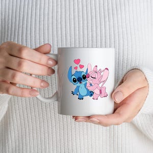 May include: White ceramic mug featuring a blue and pink cartoon couple embracing, with pink hearts floating above them. The mug has a curved handle and is held by a person wearing a white sweater.
