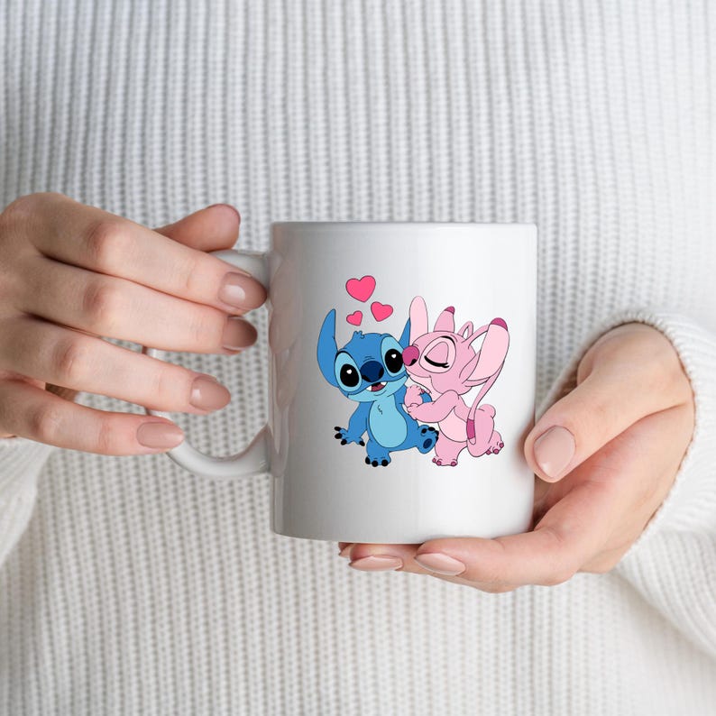 May include: White ceramic mug featuring a blue and pink cartoon couple embracing, with pink hearts floating above them. The mug has a curved handle and is held by a person wearing a white sweater.