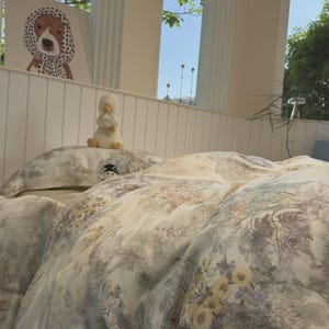 May include: A bed set featuring a white duvet cover and pillowcases with a detailed botanical print in shades of yellow, green, and grey. A small plush duck toy sits on the bed, and a framed illustration of a dog in a polka dot headscarf is visible.