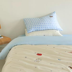 May include: A bedding set featuring a light blue and white gingham pillowcase with a small embroidered dog, a plain cream pillowcase, and a cream duvet cover decorated with small grey dots and embroidered icons of a red apple and dice.