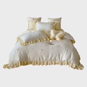 May include: A pale yellow bedding set featuring a duvet cover and multiple pillows with ruffled edges. The fabric has a smooth finish with small embroidered floral details. A butterfly-shaped decorative pillow is centred on the bed.