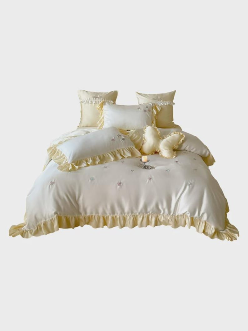 May include: A pale yellow bedding set featuring a duvet cover and multiple pillows with ruffled edges. The fabric has a smooth finish with small embroidered floral details. A butterfly-shaped decorative pillow is centred on the bed.