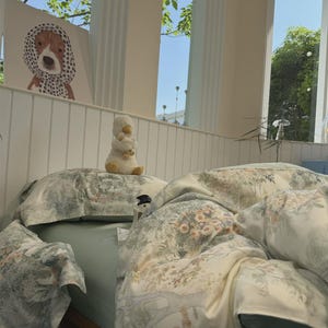 May include: A bed featuring a light green fitted sheet and a duvet cover with a detailed botanical print of trees, flowers, and foliage in muted tones. A plush white duck toy sits on the pillow, and a framed illustration of a dog is nearby.