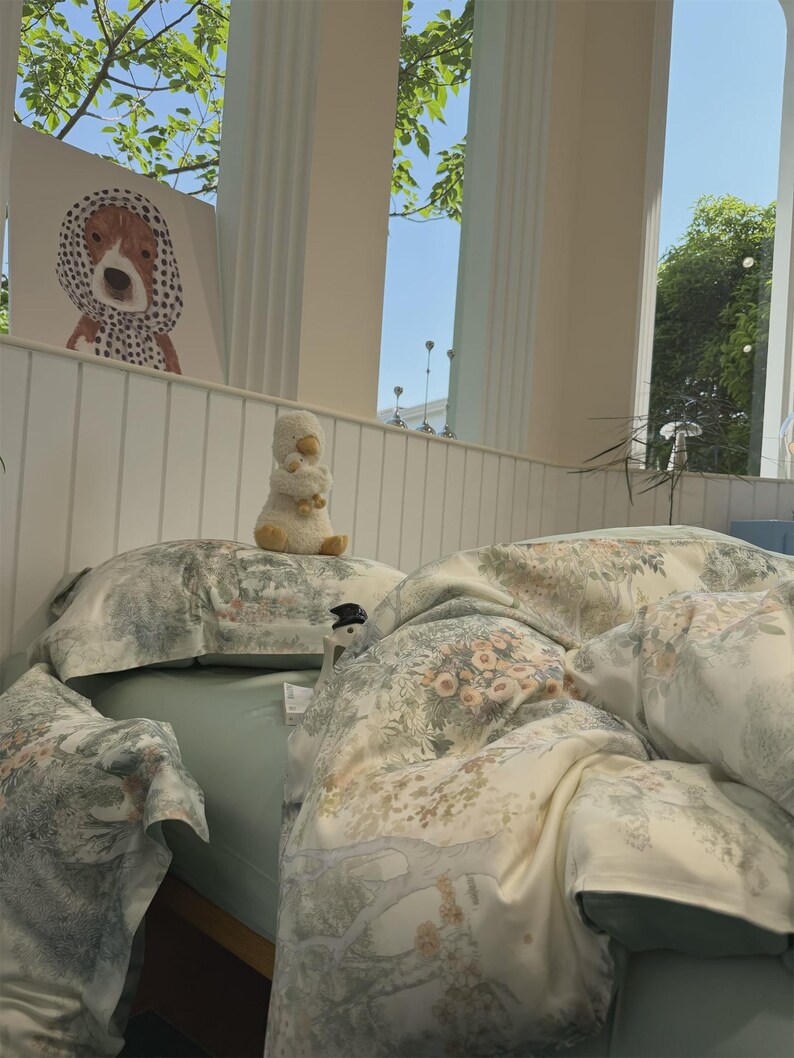 May include: A bed featuring a light green fitted sheet and a duvet cover with a detailed botanical print of trees, flowers, and foliage in muted tones. A plush white duck toy sits on the pillow, and a framed illustration of a dog is nearby.