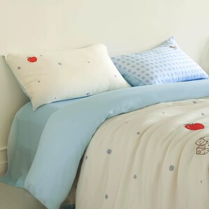 May include: A bedding set featuring a cream-colored duvet cover and pillowcase with a pattern of small gray dots and red apple illustrations. A solid light blue flat sheet and a second pillowcase with a light blue and white checkered pattern.
