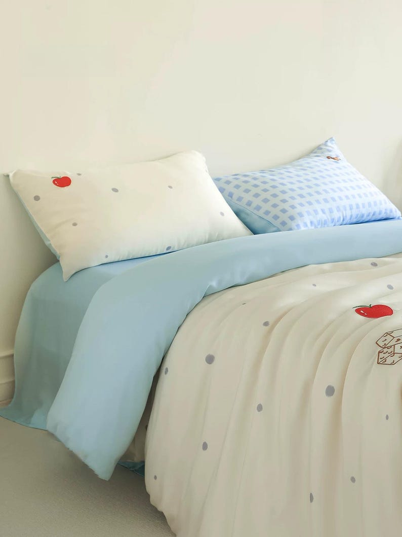 May include: A bedding set featuring a cream-colored duvet cover and pillowcase with a pattern of small gray dots and red apple illustrations. A solid light blue flat sheet and a second pillowcase with a light blue and white checkered pattern.