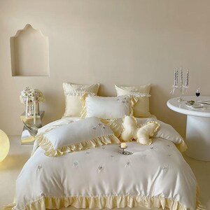 May include: A white bedding set featuring a soft yellow ruffled trim, including a duvet cover, two square pillows, two rectangular pillows, and a butterfly-shaped decorative pillow. The fabric is adorned with small, embroidered butterfly motifs.