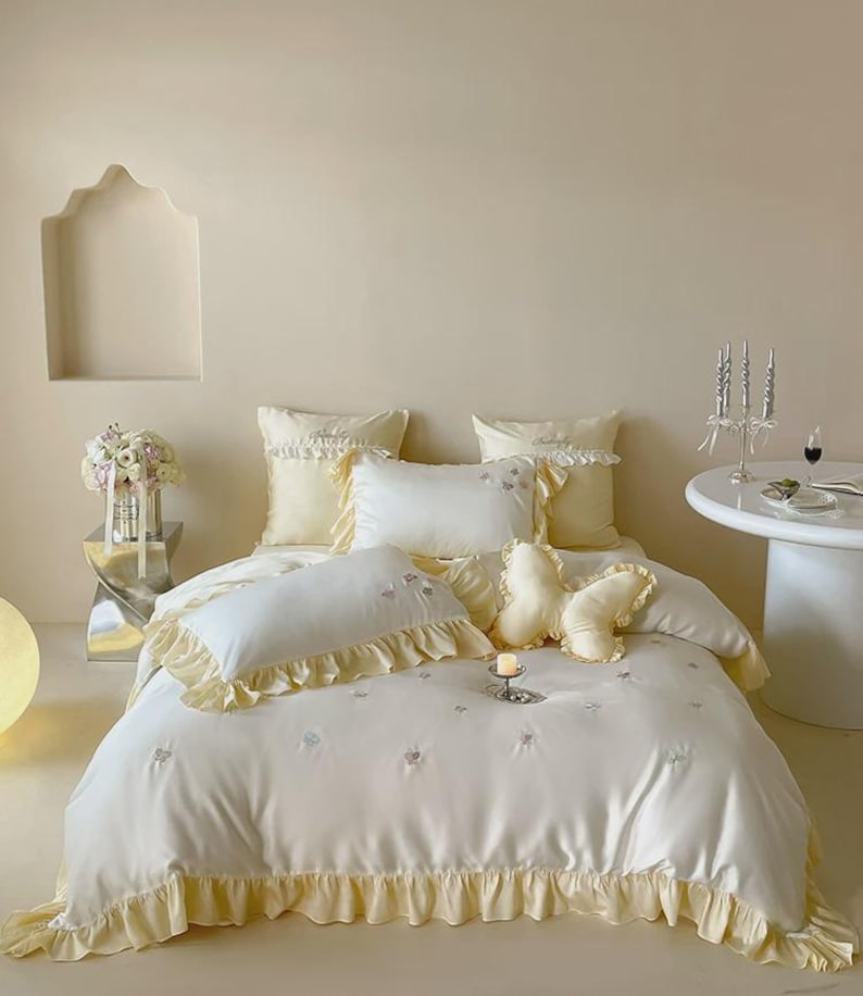 May include: A white bedding set featuring a soft yellow ruffled trim, including a duvet cover, two square pillows, two rectangular pillows, and a butterfly-shaped decorative pillow. The fabric is adorned with small, embroidered butterfly motifs.