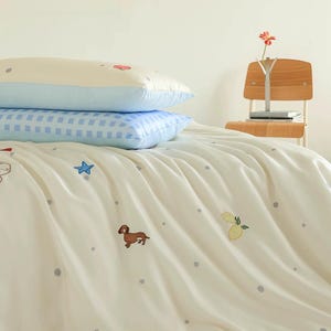 May include: A bedding set featuring a cream-colored duvet cover with small grey polka dots, embroidered brown dachshunds, blue stars, and yellow lemons. Two stacked pillows are included: one cream-colored and one with a blue and white gingham pattern.