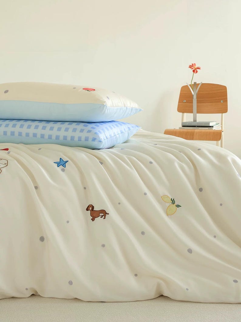 May include: A bedding set featuring a cream-colored duvet cover with small grey polka dots, embroidered brown dachshunds, blue stars, and yellow lemons. Two stacked pillows are included: one cream-colored and one with a blue and white gingham pattern.