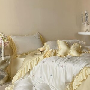 May include: A bedding set featuring a white duvet cover and pillowcases with pale yellow ruffled edges. The duvet cover is decorated with small embroidered butterflies. A butterfly-shaped decorative pillow in pale yellow sits on the bed.