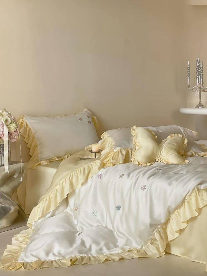May include: A bedding set featuring a white duvet cover and pillowcases with pale yellow ruffled edges. The duvet cover is decorated with small embroidered butterflies. A butterfly-shaped decorative pillow in pale yellow sits on the bed.