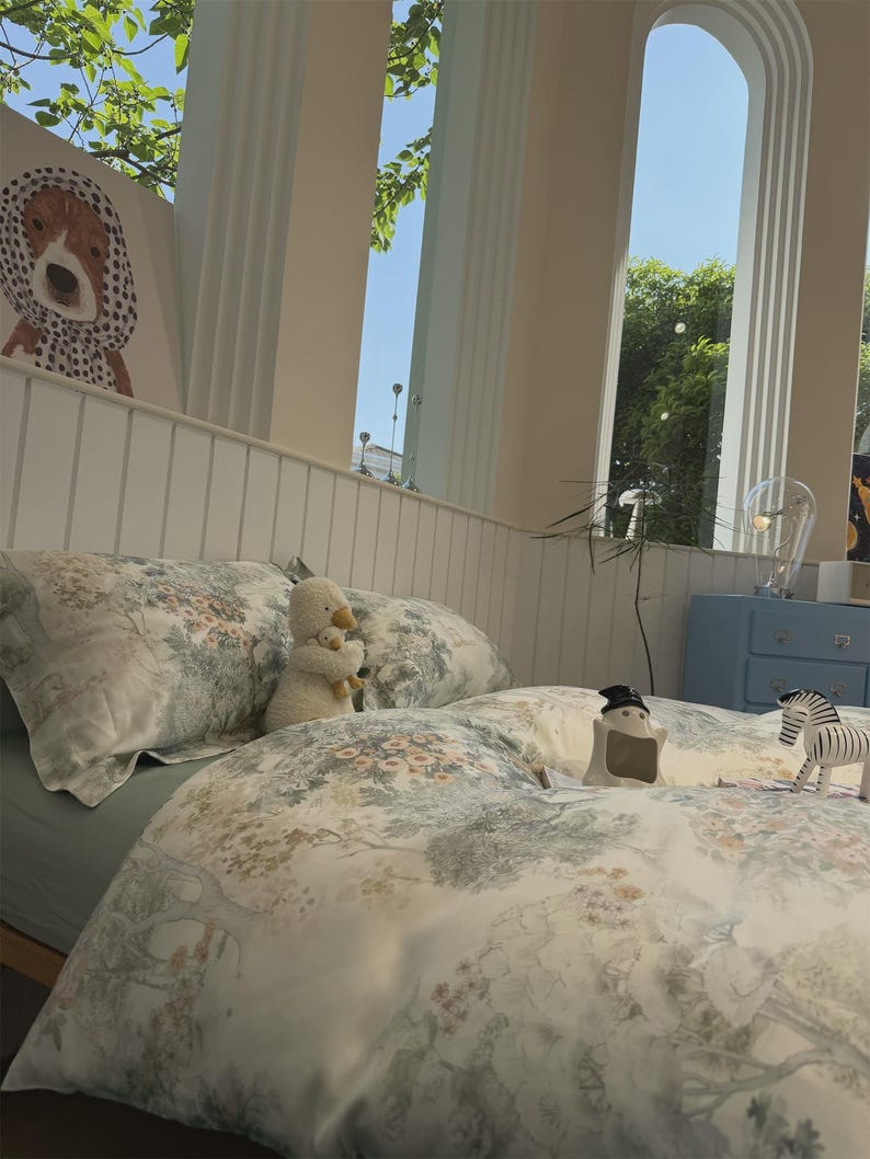 May include: A bedding set featuring a light-colored duvet cover and pillowcases with a detailed botanical print of trees, flowers, and foliage in muted green, blue, and orange tones. The set is displayed on a bed with a plush white duck toy.