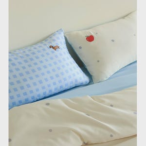 May include: Two rectangular pillows and a duvet cover. One pillow features a light blue and white gingham pattern with a small brown dog embroidery. The other pillow and the duvet cover are cream-colored with small grey polka dots and a red apple.