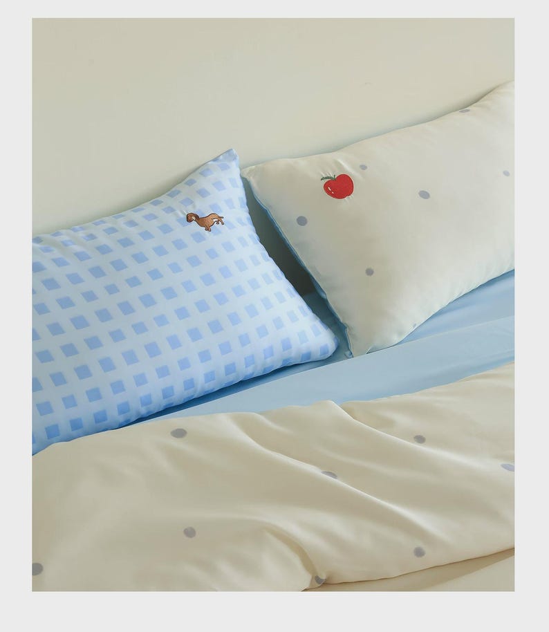 May include: Two rectangular pillows and a duvet cover. One pillow features a light blue and white gingham pattern with a small brown dog embroidery. The other pillow and the duvet cover are cream-colored with small grey polka dots and a red apple.