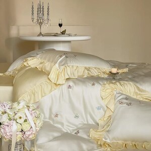 May include: A cream-coloured bedding set featuring a duvet cover and pillowcases with ruffled edges in a pale yellow hue. The fabric is adorned with small, embroidered butterfly motifs in various pastel colours scattered across the surface.