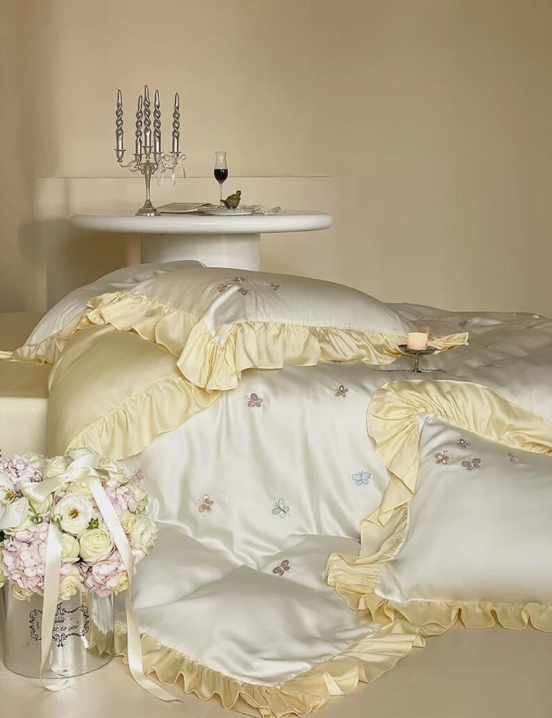 May include: A cream-coloured bedding set featuring a duvet cover and pillowcases with ruffled edges in a pale yellow hue. The fabric is adorned with small, embroidered butterfly motifs in various pastel colours scattered across the surface.