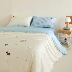 May include: A bedding set featuring a cream-colored duvet cover with small gray polka dots and embroidered motifs of a brown dachshund, a blue star, a lemon, and playing cards. It includes one cream pillowcase and one blue gingham pillowcase.