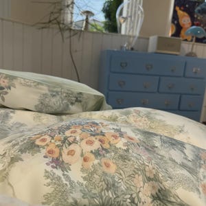 May include: A cream-colored duvet cover and pillowcase set featuring a repeating pattern of soft peach-colored flowers, green foliage, and intricate botanical illustrations. The fabric has a smooth texture with a detailed floral print throughout.