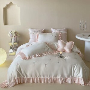 May include: A white bedding set featuring light pink ruffled edges and embroidered butterfly accents. The set includes a duvet cover, two rectangular pillows with ruffles, and a decorative butterfly-shaped pillow in soft pink fabric.