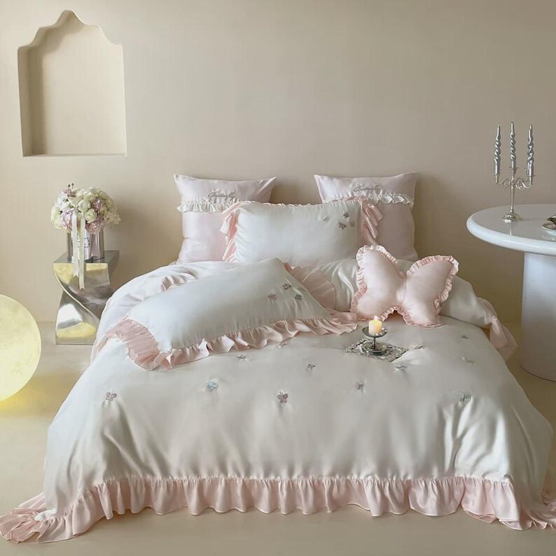 May include: A white bedding set featuring light pink ruffled edges and embroidered butterfly accents. The set includes a duvet cover, two rectangular pillows with ruffles, and a decorative butterfly-shaped pillow in soft pink fabric.
