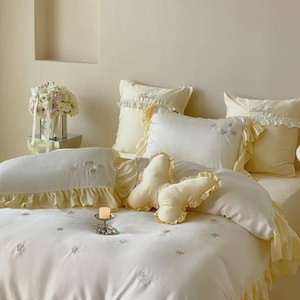 May include: A cream-coloured bedding set featuring a duvet cover and pillowcases with ruffled edges and embroidered butterfly motifs. A butterfly-shaped throw pillow sits in the centre of the bed, which is adorned with small floral patterns.