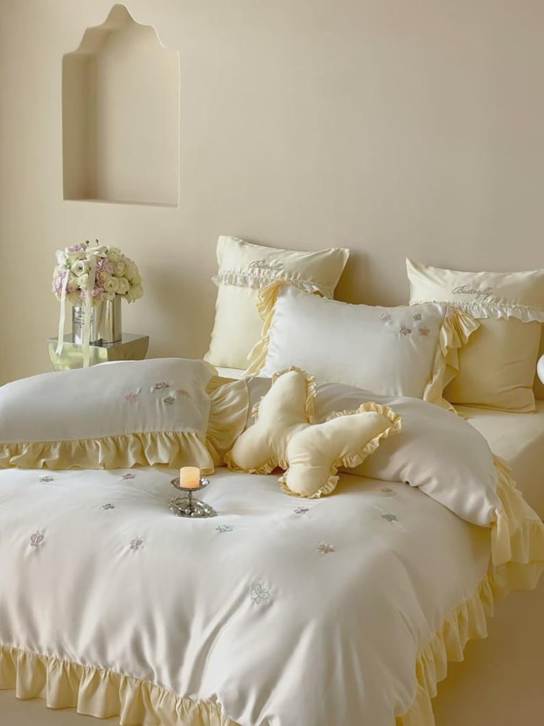 May include: A cream-coloured bedding set featuring a duvet cover and pillowcases with ruffled edges and embroidered butterfly motifs. A butterfly-shaped throw pillow sits in the centre of the bed, which is adorned with small floral patterns.