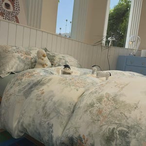 May include: A bedding set featuring a cream-colored duvet cover and pillowcases with a detailed green and muted orange botanical print of trees, flowers, and foliage. Small plush animal figures are placed on top of the bedspread.