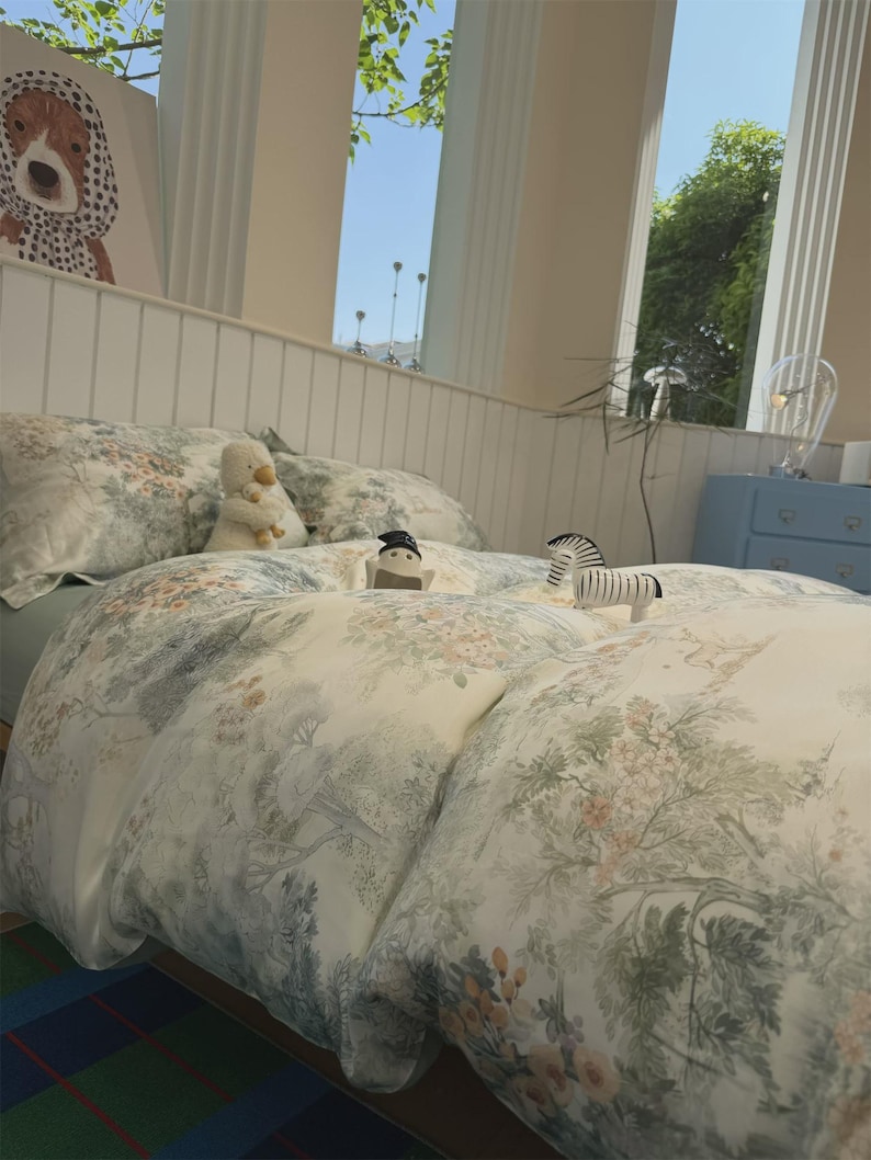 May include: A bedding set featuring a cream-colored duvet cover and pillowcases with a detailed green and muted orange botanical print of trees, flowers, and foliage. Small plush animal figures are placed on top of the bedspread.