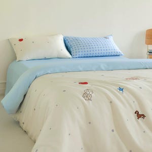 May include: A bedding set featuring a cream-colored duvet cover and pillowcase with small grey polka dots and embroidered motifs including a red apple, white dice, a blue star, and a brown dachshund. A second pillowcase has a blue gingham pattern.
