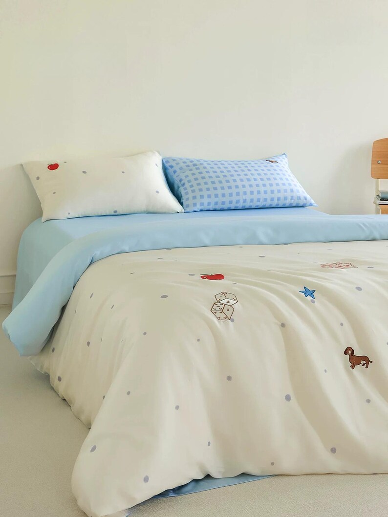 May include: A bedding set featuring a cream-colored duvet cover and pillowcase with small grey polka dots and embroidered motifs including a red apple, white dice, a blue star, and a brown dachshund. A second pillowcase has a blue gingham pattern.