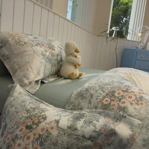 May include: A bed set featuring a light green fitted sheet and pillows with a white, green, and peach floral toile pattern. A plush white duck toy holding a smaller duckling sits upright on the bed between the pillows.