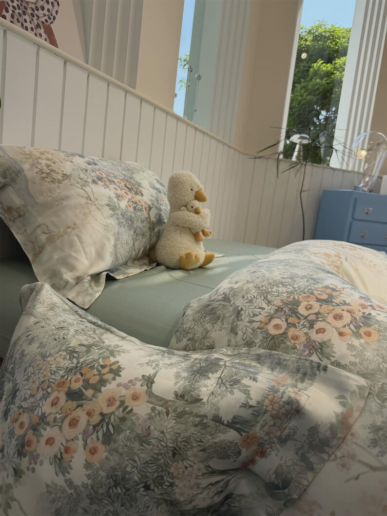 May include: A bed set featuring a light green fitted sheet and pillows with a white, green, and peach floral toile pattern. A plush white duck toy holding a smaller duckling sits upright on the bed between the pillows.
