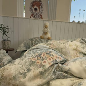 May include: A bedding set featuring a soft cream background with a detailed botanical print in muted shades of sage green, dusty rose, and soft yellow. The pattern includes clusters of flowers, foliage, and trees, creating a layered visual effect.