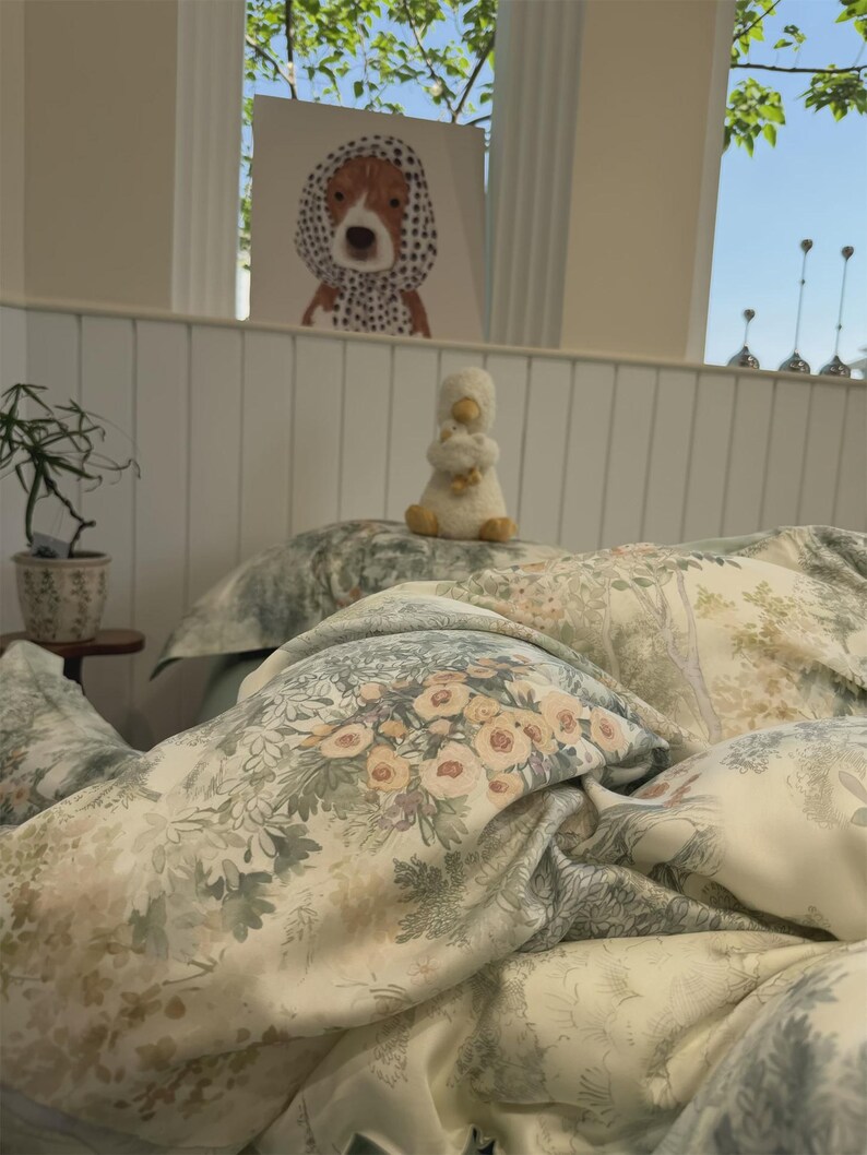 May include: A bedding set featuring a soft cream background with a detailed botanical print in muted shades of sage green, dusty rose, and soft yellow. The pattern includes clusters of flowers, foliage, and trees, creating a layered visual effect.
