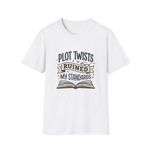 Puede incluir: Camiseta blanca con el texto "PLOT TWISTS RUINED MY STANDARDS" en un diseño caprichoso. El gráfico incluye un libro abierto, gafas y elementos decorativos. Un artículo de ropa divertido y de temática literaria.