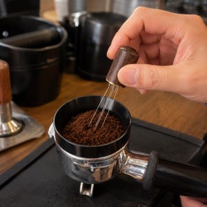 May include: A coffee distribution tool with a dark wood handle and thin metal needles is used to evenly distribute coffee grounds in a portafilter. The portafilter is black and silver, filled with brown coffee grounds.