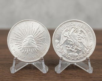 May include: Two silver Mexican coins displayed on clear plastic stands. The left coin features a Phrygian cap with rays and the text UN PESO Z 1899 F 902.7. The right coin shows an eagle perched on a cactus with the text REPUBLICA MEXICANA.