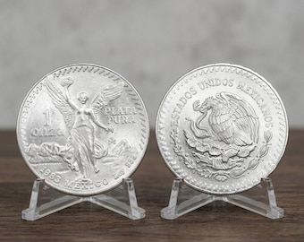 May include: Two silver Mexican Libertad coins displayed on clear plastic stands. The left coin features the Winged Victory statue, the text 1 ONZA, PLATA PURA, 1983, and MEXICO. The right coin shows the Mexican coat of arms and ESTADOS UNIDOS MEXICANOS.