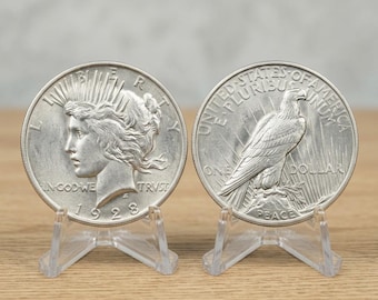 May include: Two silver-toned Peace dollar coins dated 1928, each displayed on a clear plastic stand. The left coin shows the profile of Liberty with the text LIBERTY, IN GOD WE TRUST, and 1928. The right coin features an eagle with the text UNITED STATES OF AMERICA, E PLURIBUS UNUM, ONE DOLLAR, and PEACE.