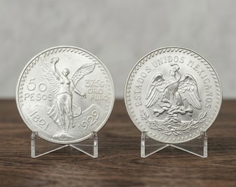 May include: Two silver-colored coins displayed on clear plastic stands. The left coin features a winged figure, the text 50 PESOS, 37.5gr ORO PURO, and the years 1821 and 1929. The right coin shows an eagle perched on a cactus with ESTADOS UNIDOS MEXICANOS.