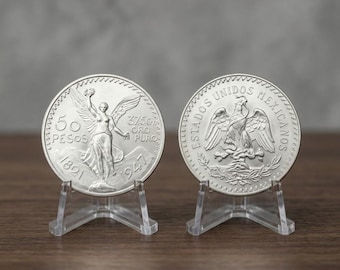 May include: Two silver-colored Mexican 50 Pesos coins displayed on clear plastic stands. The left coin features a winged figure, the text 50 PESOS, 37.5GR ORO PURO, and the years 1821 and 1947. The right coin shows an eagle and ESTADOS UNIDOS MEXICANOS.
