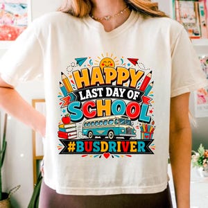 Puede incluir: Una camiseta de color crema con un gráfico colorido y el texto "HAPPY LAST DAY OF SCHOOL #BUSDRIVER". El diseño incluye un sol amarillo, lápices, una pila de libros con una manzana y un autobús escolar azul rodeado de pequeñas estrellas.