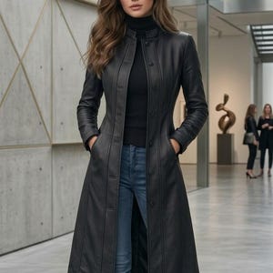 May include: A long black leather coat featuring a high collar, a front button closure, and two side pockets. The coat is worn over a black turtleneck sweater, blue denim jeans, and black leather ankle boots, creating a structured silhouette.