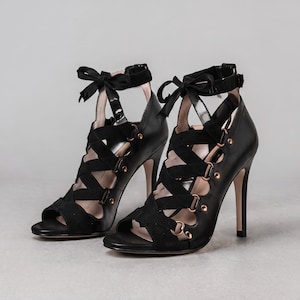 May include: A pair of black high-heeled sandals featuring a crisscross strap design across the front, rose gold-toned metal rings, and an ankle strap that ties into a bow. The shoes have an open-toe construction and a slim stiletto heel.