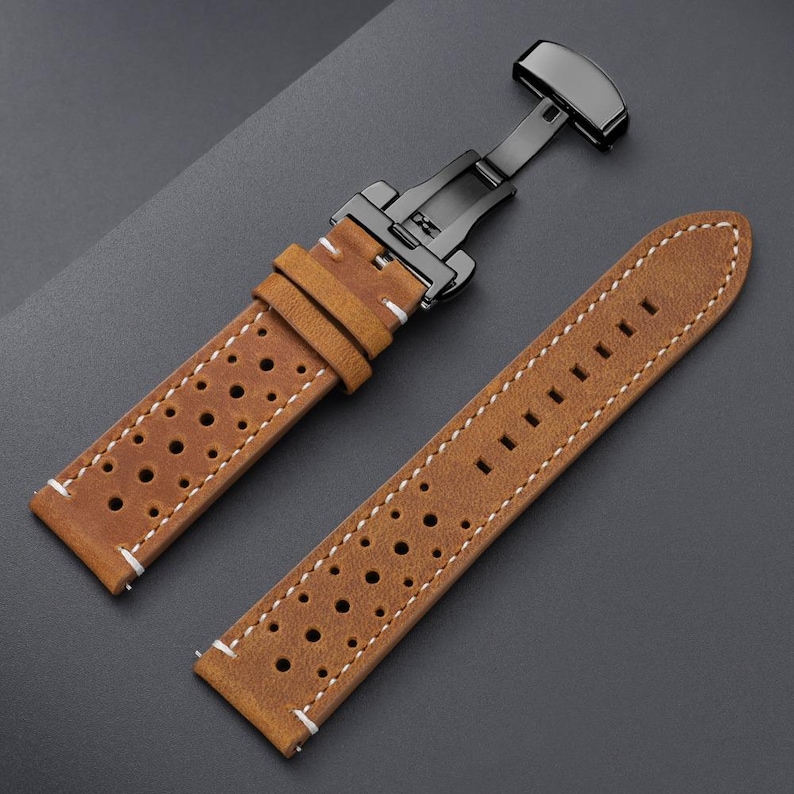 May include: A light brown leather watch strap with a black clasp. The strap has a perforated design and white stitching. The watch band is shown on a dark gray surface.