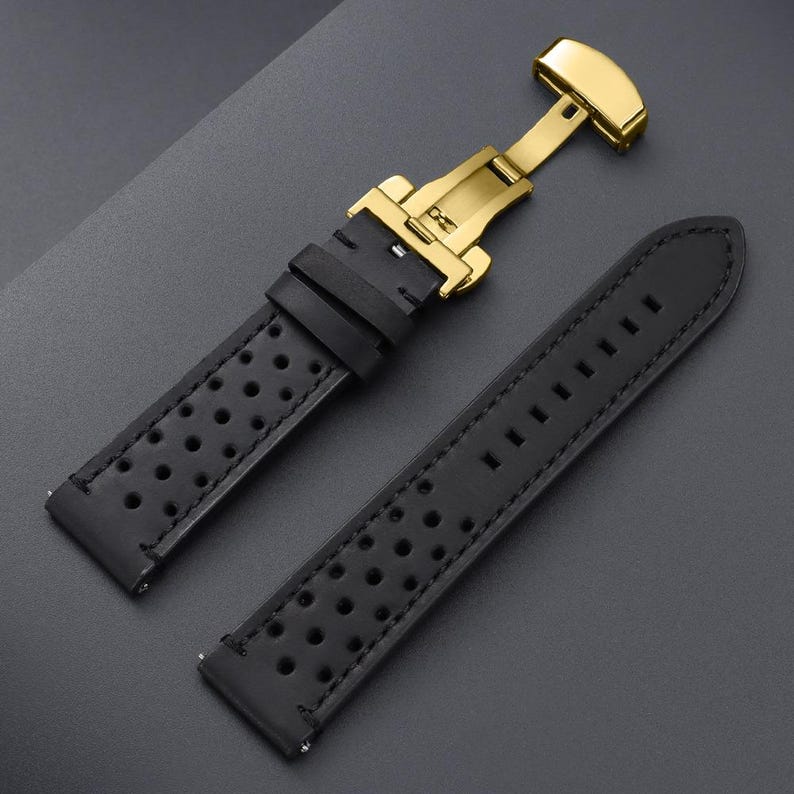 May include: Black leather watch strap with a gold-colored clasp. The strap has a perforated design and is stitched with black thread. The clasp is a butterfly style, and the strap is shown against a dark gray background.