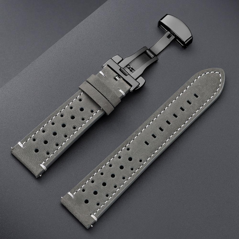 May include: Gray watch strap with white stitching and a black clasp. The strap has a perforated design and is made of a suede-like material. The watch band is shown on a dark gray surface.