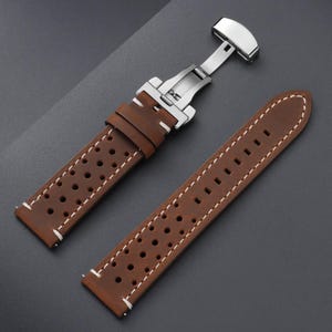 May include: Brown leather watch strap with white stitching and a stainless steel clasp. The strap features a perforated design and is shown open, displaying the clasp mechanism. The watch band is designed for a classic look.